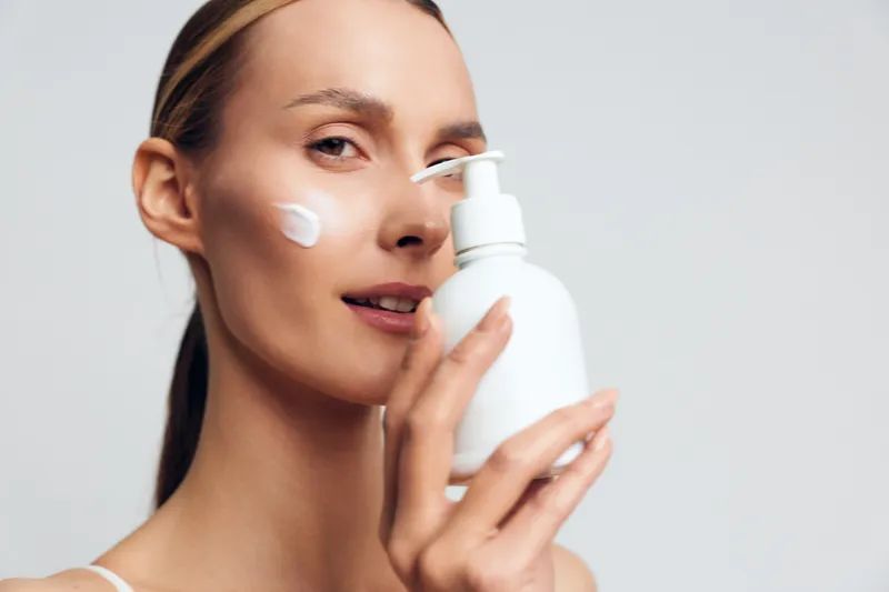 Young woman applying face cream with smooth skin holding white lotion bottle close to her face in bright studio with neutral background and natural makeup