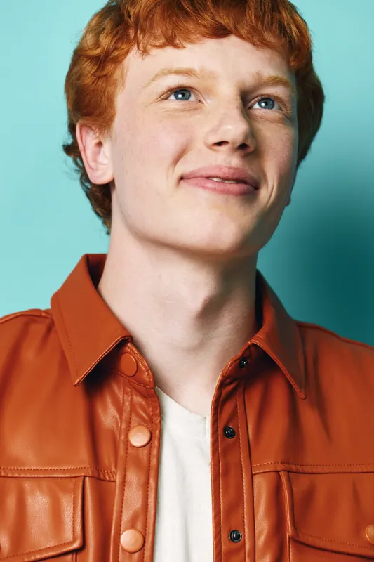 Redhead man portrait with blue eyes smiling and looking up wearing brown leather jacket and white shirt on turquoise background with soft lighting and positive expression.