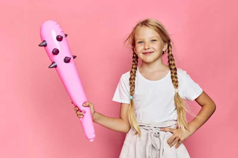 Smiling young girl with braided hair holds a pink inflatable bat with spikes against a pink background. She wears a white t-shirt and light skirt. Studio shot with playful mood.