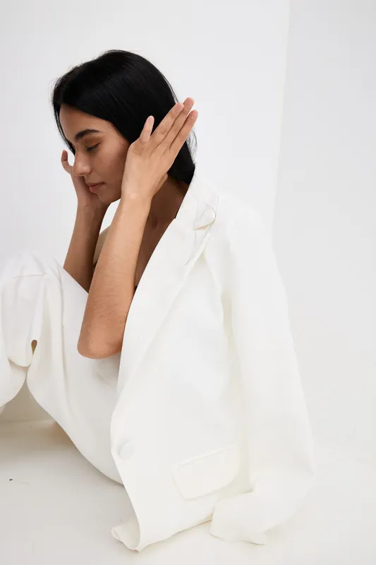 Young woman in white oversized blazer sitting thoughtfully against a minimalist white background, showcasing modern fashion and serene mood in soft natural lighting.