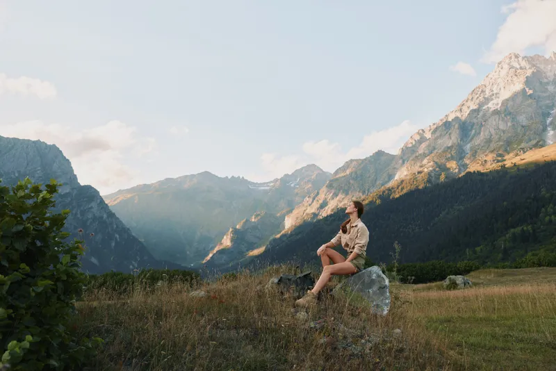 Young woman in casual clothes sitting on a rock in a mountain valley, enjoying nature and fresh air during a sunny day. Concept of lifestyle, adventure, and relaxation.