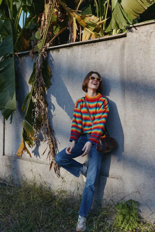 Woman in a colorful striped sweater and sunglasses leaning against a concrete wall with banana leaves, smiling and laughing in blue jeans with a casual crossbody bag in sunlit outdoor lifestyle.