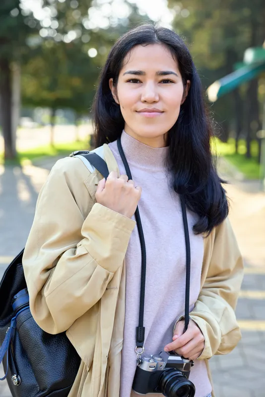 Female traveler with backpack and camera in a sunlit park, casual jacket and sweater, exploring outdoors, capturing moments, photography scene, daytime leisure and outdoor curiosity
