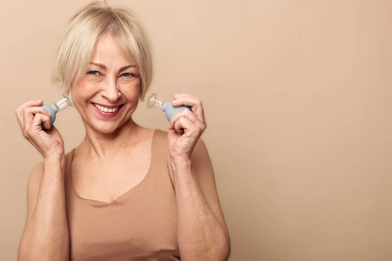 Smiling middle aged woman with short blonde hair uses facial rollers on beige background. Concept of skin care, anti aging, wellness, beauty routine, natural face massage, self care.
