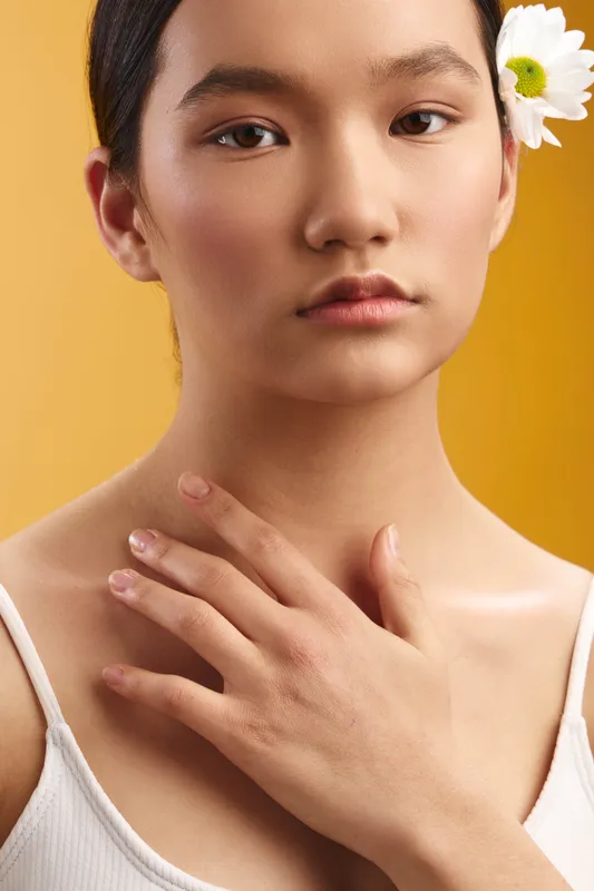 Asian woman gazes softly at the camera, a white flower tucked behind her ear. The minimalist portrait highlights serene beauty against a warm, simple backdrop.