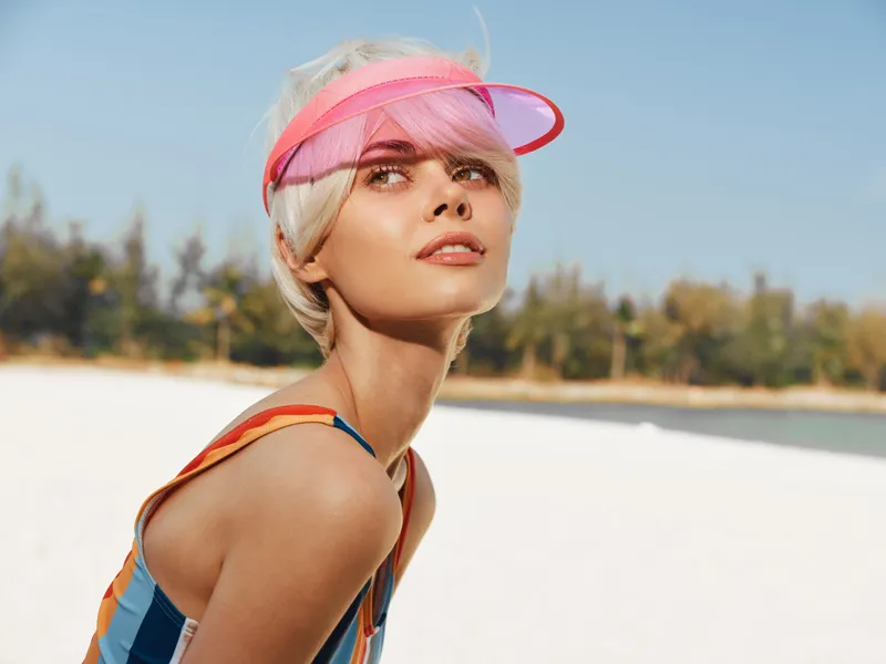 A stylish woman on a sunny beach wears a pink visor and colorful striped swimsuit, posing with a confident gaze as radiant light highlights her features.