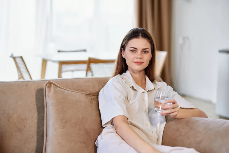 Woman portrait on a sofa in the living room holding a glass of water, relaxed at home. Indoor lifestyle scene with casual clothing, natural light and cozy modern interior.