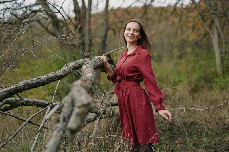 A candid environmental portrait of an adult woman in a red dress standing by a fallen tree in woodland, conveying authenticity through a genuine relaxed smile and natural light mood.