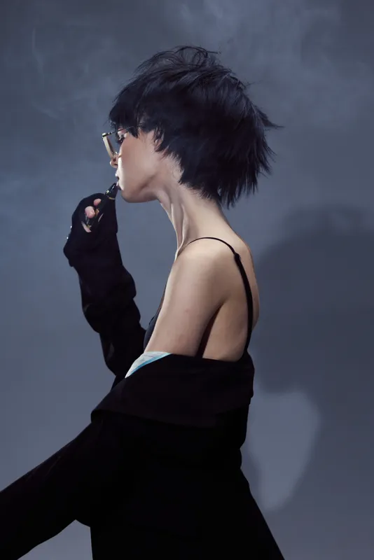 Stylish woman with short hair in dark clothes holding a lighter, moody portrait with dramatic lighting and gray background.