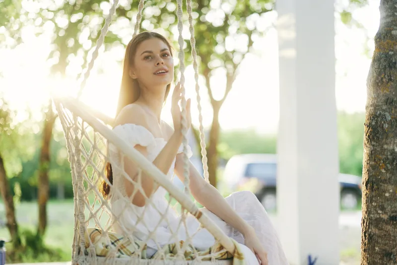 Young woman sitting on a swing in a bright garden, wearing a white dress, radiating joy and tranquility during a sunny day, perfect for summer lifestyle campaigns