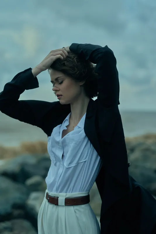 Coastline fashion shot featuring a wind swept woman in a blazer and blouse, standing by rocks with an ocean backdrop, portraying elegance, confidence and modern outdoor style
