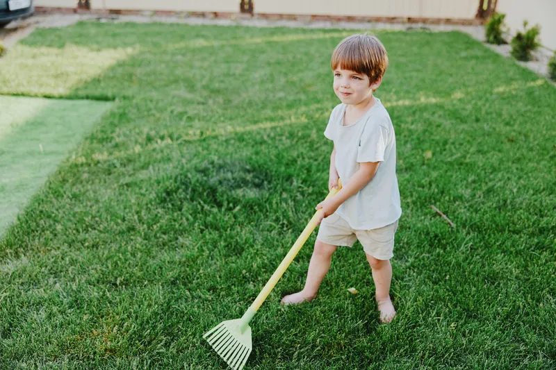Young boy with rake enjoying outdoor activities in a sunny green garden, embodying the joy of playfulness and childhood wonder during sunny days.