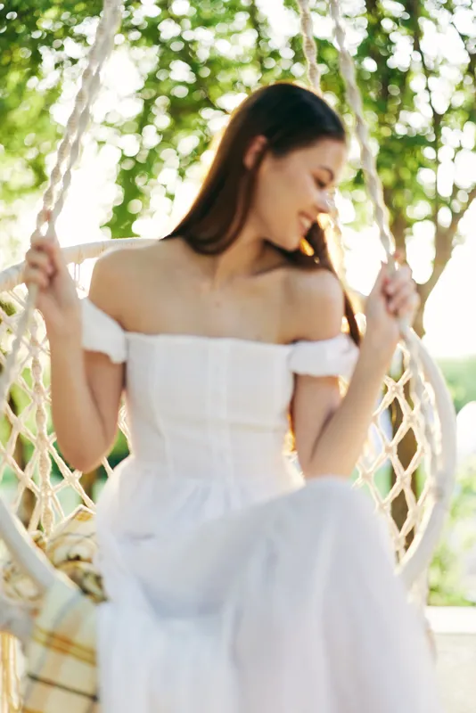 Young woman in a white dress smiling gently while sitting on a swing, surrounded by green trees and soft sunlight, conveying tranquility and femininity