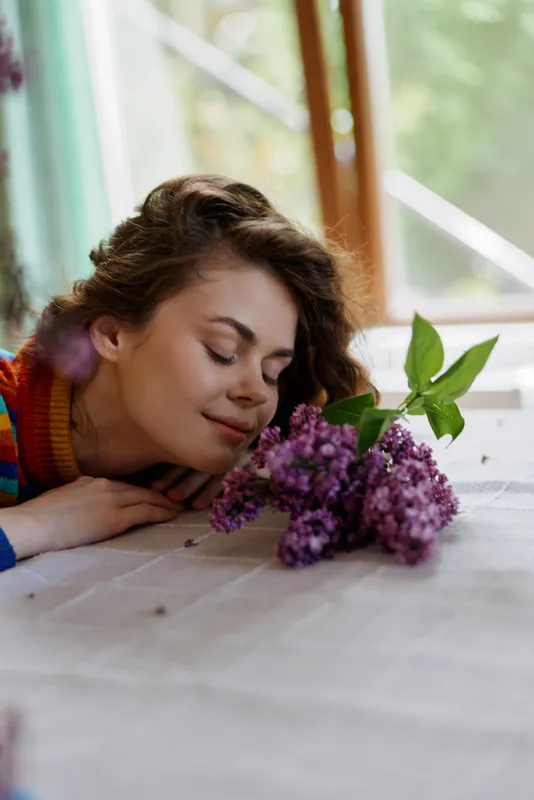 Smiling woman with brown hair resting her chin near a bouquet of lilac flowers on a wooden table. Window light creates a cozy home vibe, promoting wellness and self care.