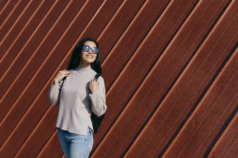 Woman in sweater and jeans posing against a diagonal wooden wall, stylish outdoor fashion, sunglasses, relaxed stance, urban vibe and natural light for lifestyle stock imagery.