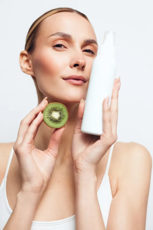 A confident young woman holds a white cosmetic bottle in one hand and a sliced kiwi fruit in the other, symbolizing natural skincare and beauty with fresh ingredients.