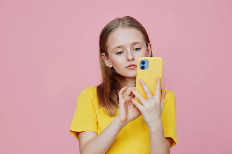 Young girl with a yellow phone, wearing a bright yellow t shirt, looking thoughtfully at the screen against a soft pink background, showcasing modern digital interaction
