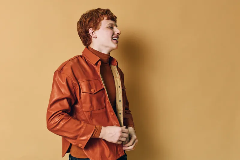 Young man with red hair wearing brown leather jacket stands against beige background smiling and looking to the side in casual style portrait.
