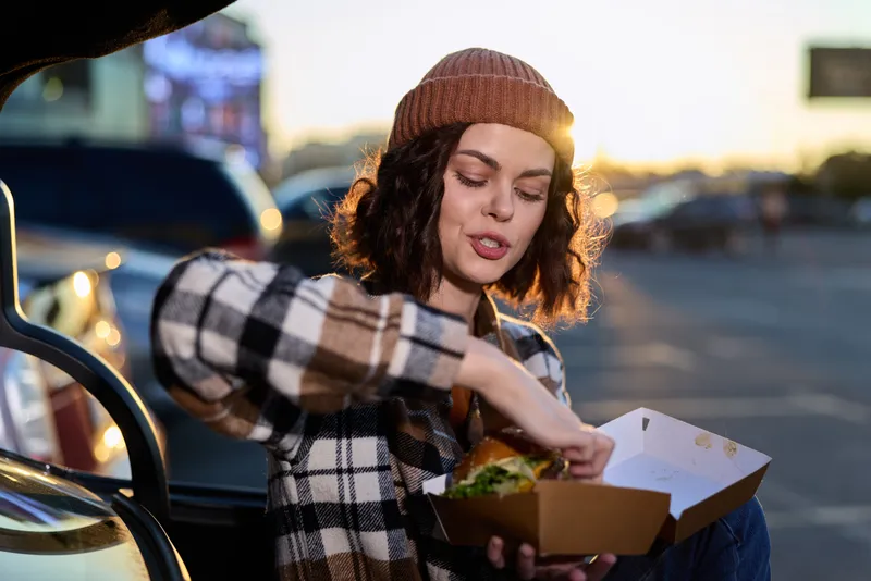 Woman eating burger from takeaway box by car trunk at golden hour, candid authenticity and emotional storytelling in a mindful living urban scene with warm golden hour glow.