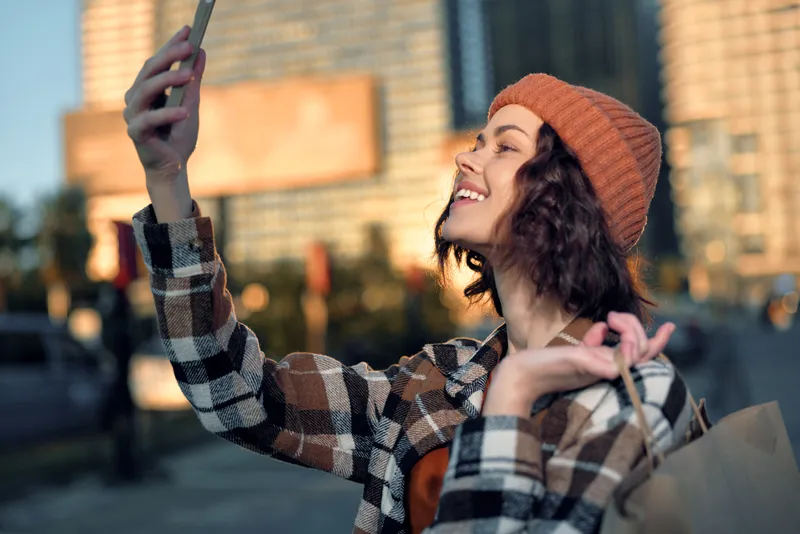 Woman taking selfie with smile in urban street, shopping bag and beanie, candid moment with golden hour glow, authenticity and emotional storytelling for mindful living lifestyle.