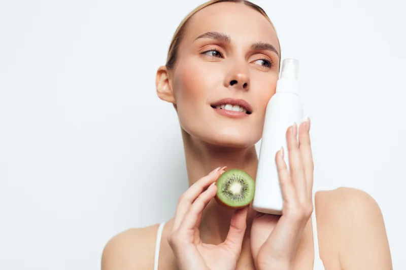 Radiant woman holding kiwi and lotion bottle, skincare routine moment, bright lighting, clean background, natural glow, hydration, relaxed expression, soft colors, fresh mood