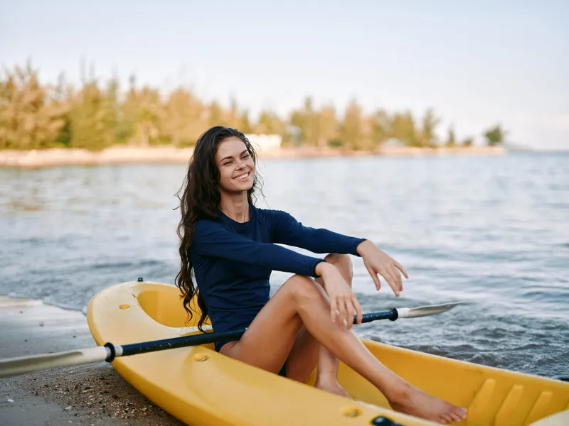 A young woman sits in a yellow kayak on a sandy shore, smiling softly as gentle waves lap the lake. Warm light and relaxed pose convey outdoor adventure.