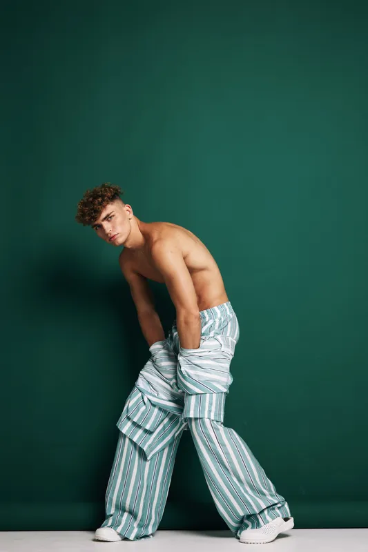 Young man with curly hair posing shirtless in oversized striped pants and white sneakers against green background in studio. Fashion, style and casual look with confident expression.