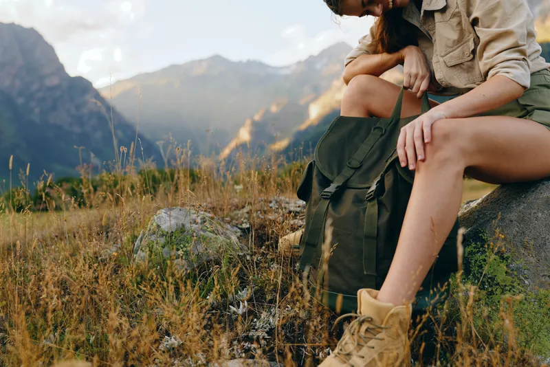 Young woman sits on a rock with a backpack in a mountain meadow, enjoying a peaceful outdoor moment during a hike.