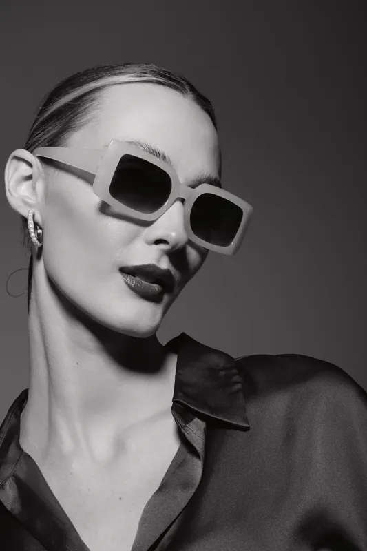 Stylish white woman with pale skin wearing large sunglasses and dark lipstick, posing in black satin blouse on dark background.