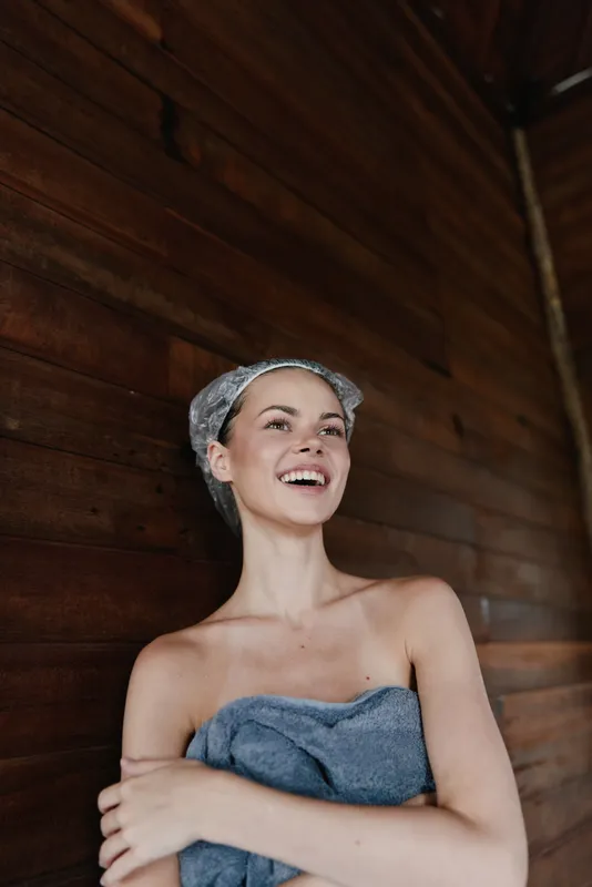 smiling woman wrapped in blue towel with wet hair towel standing against wooden wall in spa or sauna, relaxed and happy mood, natural light, wellness and self-care concept