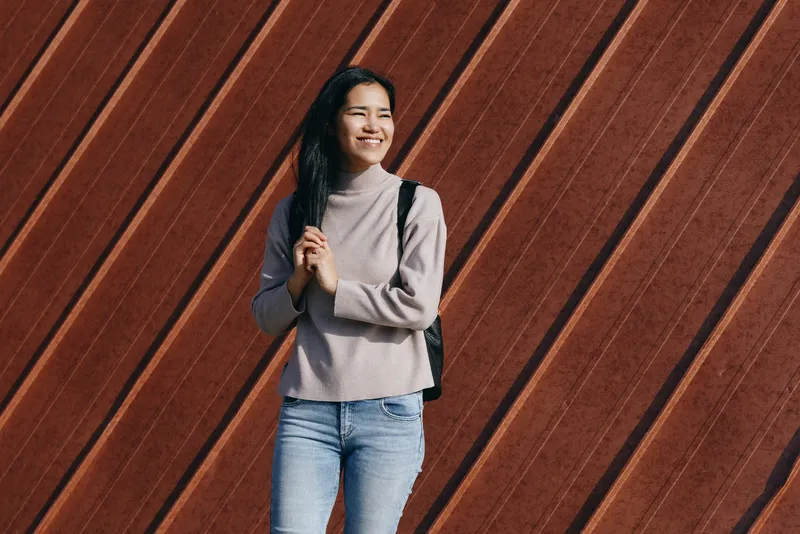 Smiling woman with backpack in casual attire stands before a diagonal wooden wall in an outdoor urban setting, conveying confident style, everyday travel mood, and friendly approach