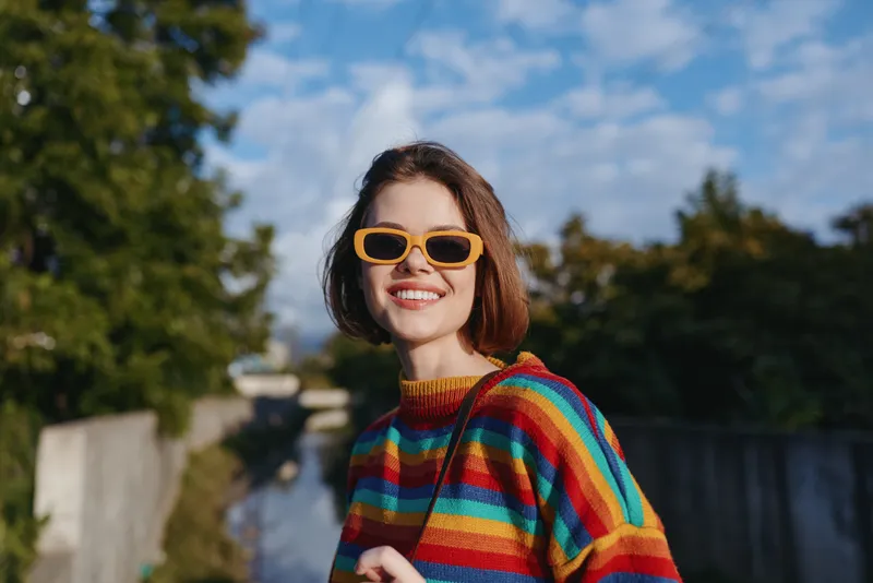 Woman smiling in orange sunglasses and rainbow sweater, portrait outdoors with blue sky, warm sunlight and shoulder bag; happy young adult in colorful casual street style, candid cheerful vibe.
