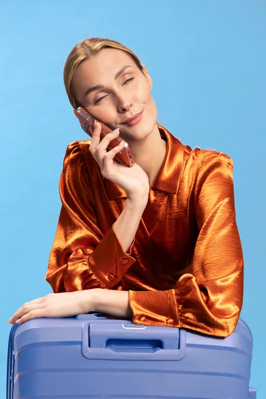 Young woman in shiny orange blouse talks on phone smiling, leaning on blue suitcase. Bright studio lighting with blue background. Concept for travel, communication, and happiness.