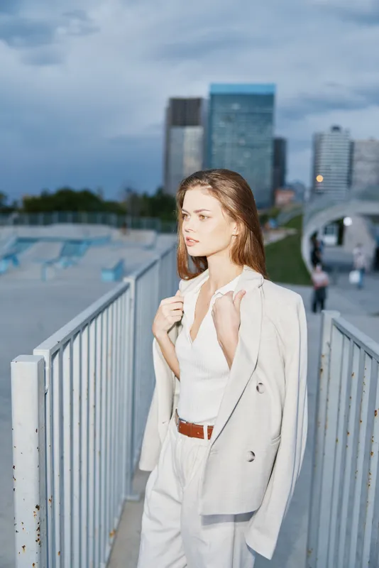 Young woman in stylish white suit walking in urban environment during cloudy day with modern buildings in background, fashion and business concept