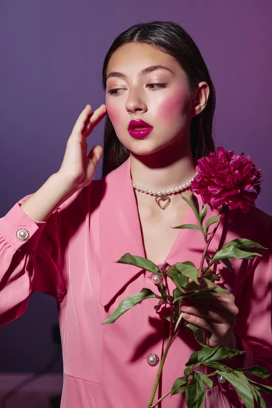 Young woman wearing bold pink makeup and glossy dark lipstick holds red flower. Studio portrait with fashion styling in a pink outfit and purple background.