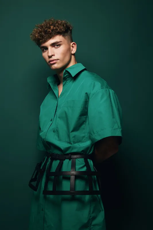 Young man with curly hair wearing green shirt dress and black belt posing against dark green background in studio portrait with serious expression and stylish look.
