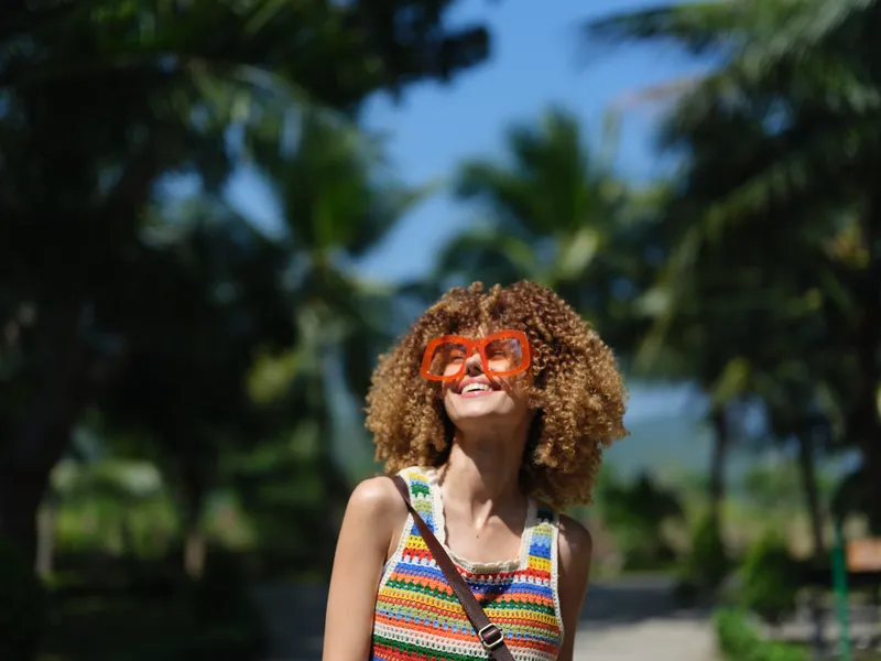 Smiling woman with curly hair wearing colorful sunglasses and summer clothes enjoying sunny tropical background and palm trees outdoors.