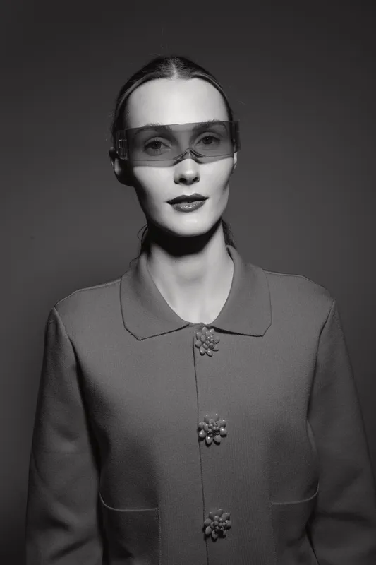 Elegant Caucasian woman with pale skin wearing futuristic sunglasses and vintage textured jacket. Studio portrait with dramatic lighting and minimalist background.