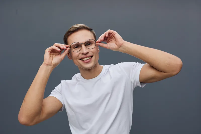 Smiling woman adjusting stylish eyeglasses while looking at camera, casual white t-shirt, exposed dark hair, cheerful expression, modern background, fashion and lifestyle concept, vibrant and candid p