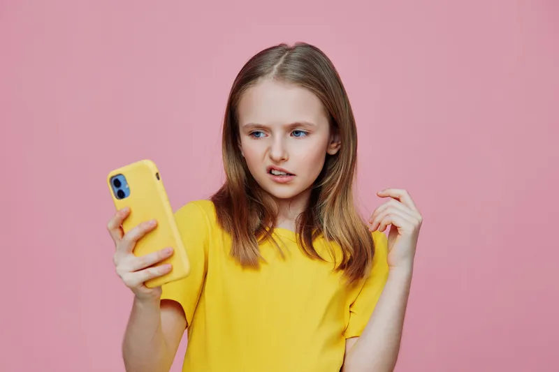 Young girl with a yellow smartphone, showing a confused expression against a pink background, representing modern communication and digital age