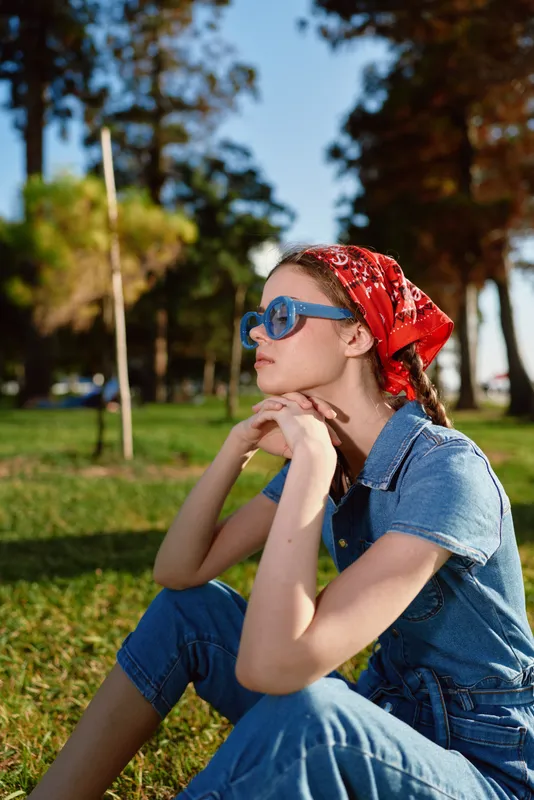 Lifestyle woman in modern boho-western denim street style sits thoughtfully outdoors wearing blue sunglasses and a red bandana under warm natural sunlight.