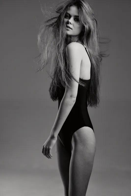 Black and white portrait of a slender woman with long hair wearing a bodysuit and posing elegantly against a plain background.