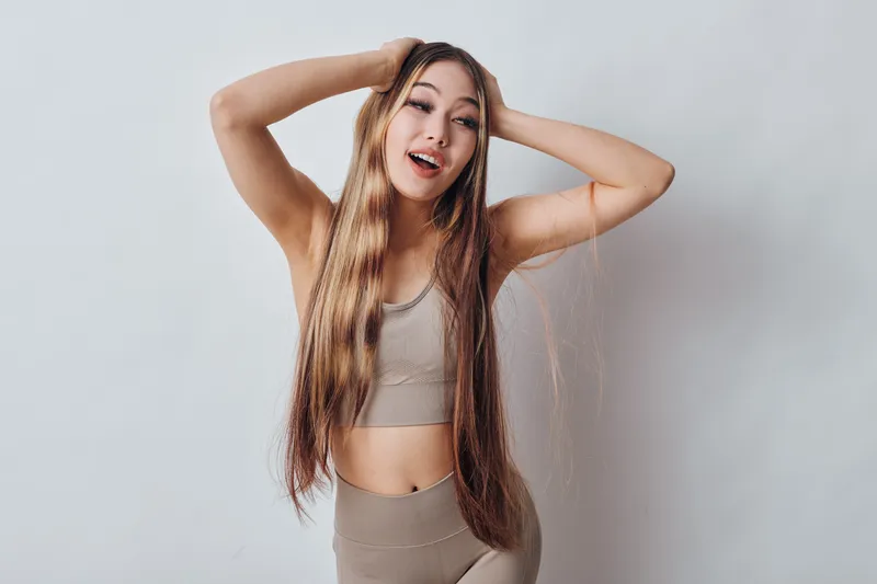 Young woman with long hair wearing beige sportswear posing with hands on head smiling and looking happy against gray background in studio. Healthy lifestyle and fitness concept.