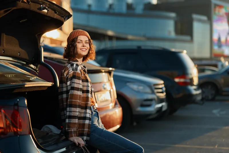 Woman leans on open car trunk in urban parking lot at golden hour, smile and candid lifestyle moment conveying authenticity, mindful living and emotional storytelling.