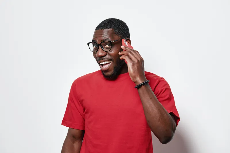 African American man with dark skin and glasses wearing red t shirt talking on smartphone with smile and excitement isolated on white background in studio portrait.