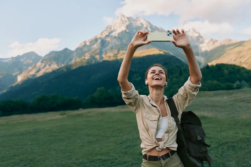 Young woman with backpack takes a cheerful selfie in a grassy field with mountain backdrop, capturing a carefree travel moment outdoors
