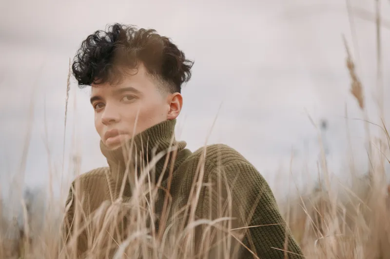 A close portrait of a young person in tall grass wearing a warm sweater, conveying authentic presence and natural expression with soft light and realistic, unposed details.