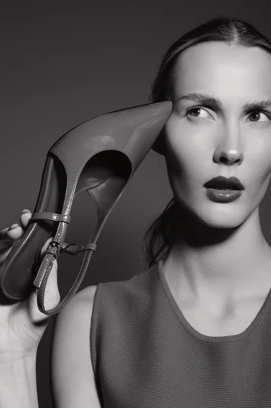 Young white woman with fair skin holds black high heel close to her face, looking sideways. Studio portrait in black and white with dramatic lighting and serious expression.
