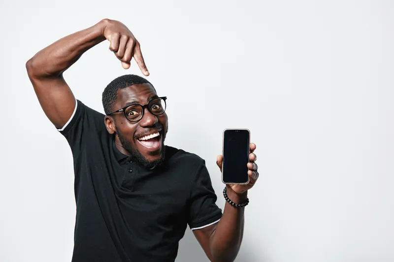 African American man with dark skin and glasses smiling and pointing at smartphone screen in black polo shirt on white background. Happy male showing mobile phone for technology or communication.