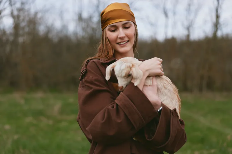 Young woman in a brown coat embraces a gentle goat kid outdoors, sharing warm affection in a rural field, with soft natural light and a calm, candid lifestyle moment.